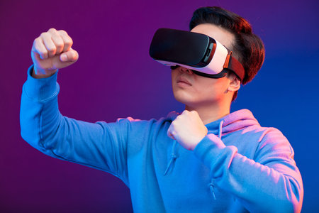 Joyful Japanese youngster playing videogame in virtual reality helmet on dark blue background. Smartphone using with virtual reality goggles. Technology, simulation, hi-tech, game conceptの写真素材
