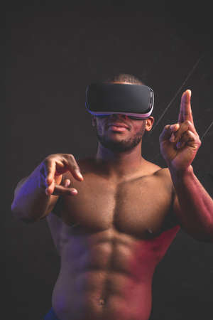 Muscular naked African boxer wearing mobile virtual reality headset prepares to fight with virtual opponent, showing gun sign against darkbackground. Sport, rivalry and virtual reality conceptの写真素材
