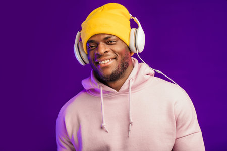 Cute African-American man in headphones dressed in street style wear, listening to music on violet...
