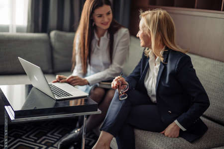 Small business concept. Young creative small business partners working with new startup project in modern office. Two young women sitting on sofa and looking at laptop, analyzing risks and benefitsの写真素材