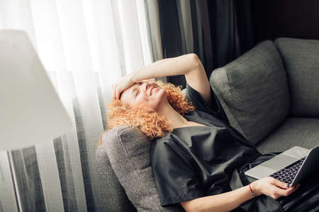 Happy relaxed woman with bushy hairstyle resting in a semi lying position on couch enjoying sun rays and good news from her boyfriend.の写真素材