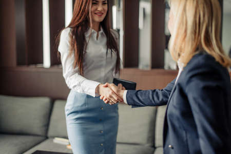 Two caucasian businesswomen in office clothes shaking hands at the meeting as they close a partnership, focus on handsの写真素材