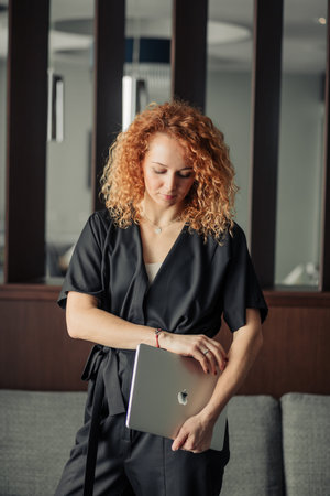 Young female fashion designer with bushy red hair dressed in trendy black overalls looking camera...