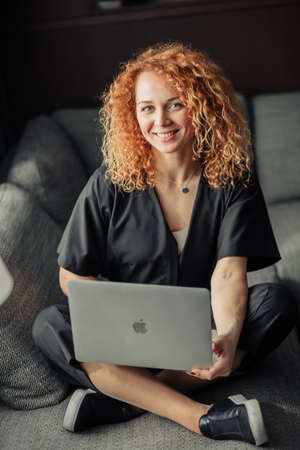 Happy smiling redheaded woman entrepreneur in startup company looking at camera while working at laptop comfortably sitting at sofa.の写真素材