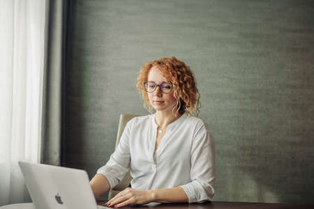 Happy smiling red-haired woman in white blouse and eyewear working on laptop sitting against grey wallの写真素材
