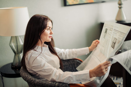 charming woman sitting in arm chair and reading newspaper in hotel room.