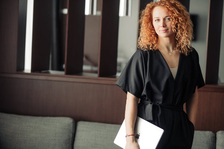 Young female fashion designer with bushy red hair dressed in trendy black overalls looking camera...