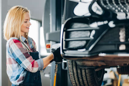 positive girl using impact wrench at workplace. side view photo. occupation, free time, unusual job for girlsの写真素材