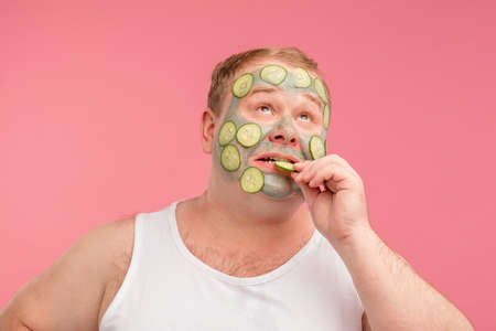 Young man in white undershirt having doubts and with confuse face expression while applying cucumber mask on his face, isolated pink backgroundの写真素材