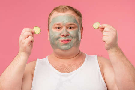 Happy middle aged man in underwear applies clay mask to cleanse the skin and cucumber slices to moisturaze, isolated on pink background. Grooming and skin care conceptの写真素材
