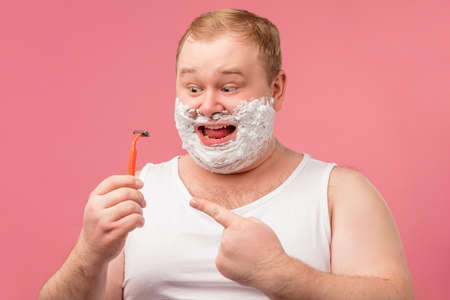 Cheerful surprised man with shaving foam on his face looking at the unknown old razor, isolated over pink background.の写真素材