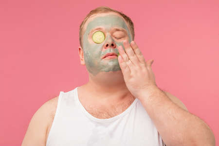 Cheerful funny man in white t-shirt has clay mask, expresses positive emotions and experiments with cucumber slices applying it on eyes and face isolated on pink wall. Beauty and spa concept.の写真素材