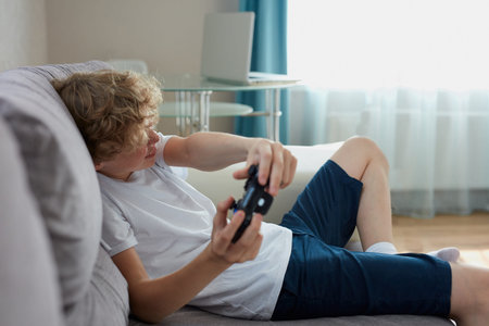 playful teenager keen on video games, sit on sofa in living room holding console in hands, enjoy,...