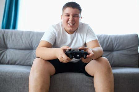 fat caucasian teenager boy enjoys video games, overweight boy sits on sofa holding console in hands, concentrated on game, look at screenの写真素材