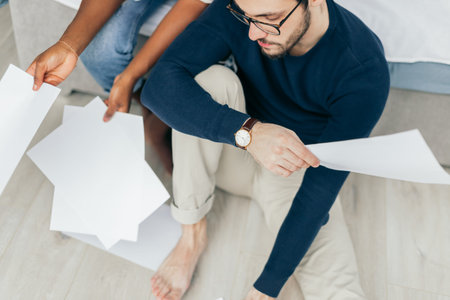 African and Caucasian mixed race married couple sitting on the floor at home checking unpaid bills,...