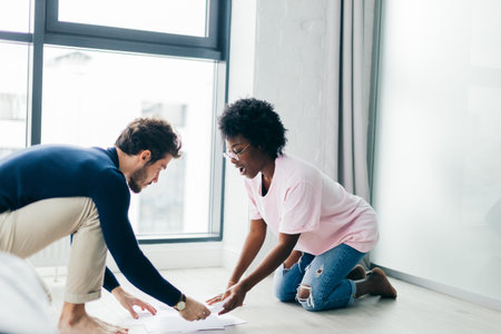 Interracial couple of young newlyweds sit together on floor of their rented appartments, busy with composing financial report for tax service, using paper documentsの写真素材