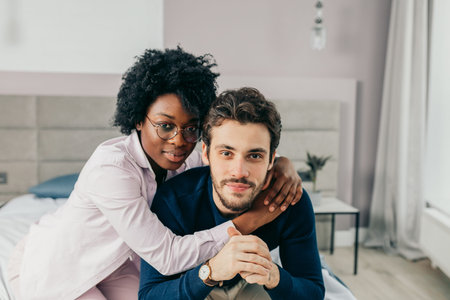 Portrait of young mixed race loving couple having romantic times in bedroom. African woman embracing affectionately her european american friend.の写真素材