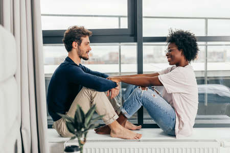Interracial married couple embrace affectionately at home interior, sitting near large window, feel support and emotional resource from each other, have close relationship.の写真素材