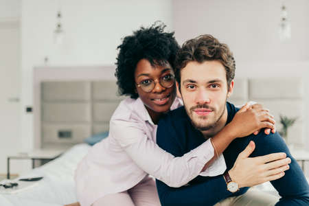Happy African american girl embracing her handsome caucasian boyfriend while sitting on bed in bedroom with modern grey colored interior and big windows.の写真素材