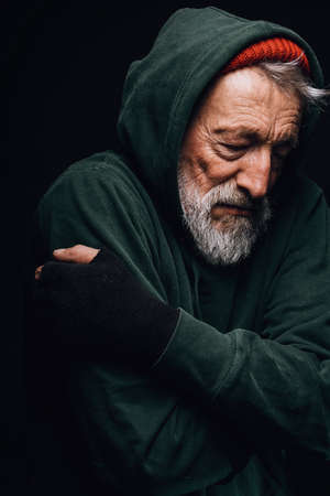 Old grey-bearded frozen tramp man with grey beard embracing himself to warm up posing over black background.の写真素材