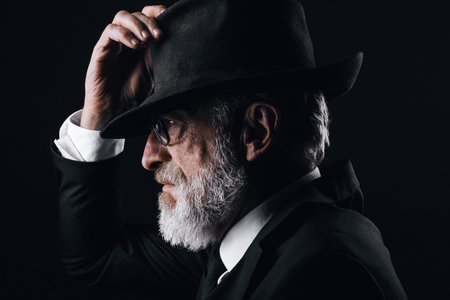 An old male actor playing an English spy role, dressed in black suit, hiding behind fedora hat, having mysterious elegant look. Isolated studio portrait over dark backgroundの写真素材
