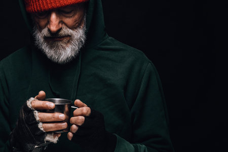 Old homeless man with grey beard covering up in green decrepit wear holding a mug of hot tea to warm...