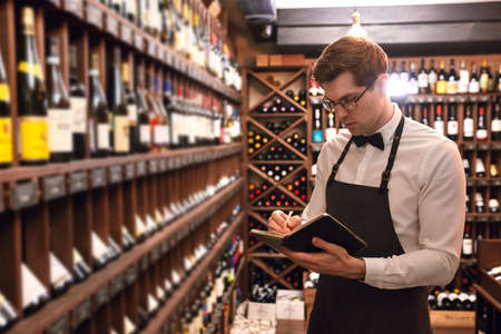Professional male cavist taking notes at degustation card standing near wine shelves of european wine boutiuqueの写真素材