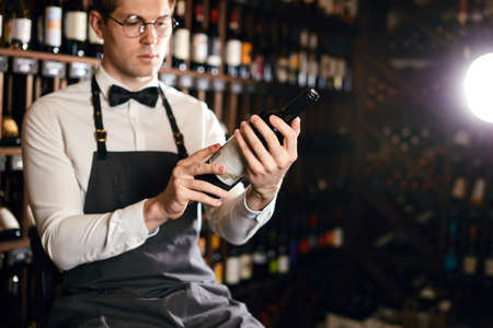 Young caucasian cavist dressed in white shirt and bowtie working in big vine shop presenting a bottle of red wine to customerの写真素材