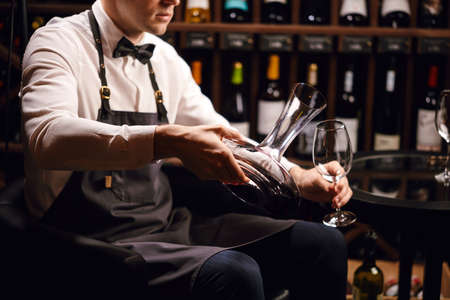 Cropped view of professional sommelier pouring red wine from bottle into decanter at table in wine restaurant.の写真素材