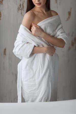 Brown-haired young woman in a bathrobe looking at camera and posing, standing turned with bare shouldersの写真素材