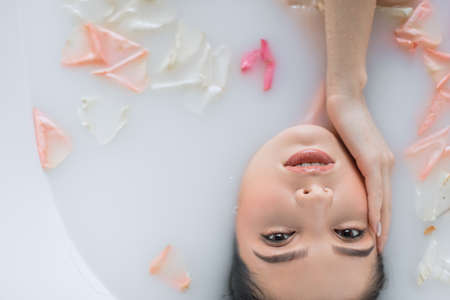 Young woman getting spa treatment at beauty salon. Female face close up, looking at camera while enjoying in milky aroma water with petals bath.の写真素材