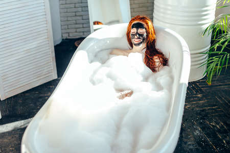 Skin care - Young lady with facial black clay mask applyed on her face relaxing in foam bath, getting rejuvenation spa and beaty procedure at homeの写真素材