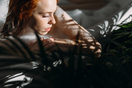 Beautiful sensual red-haired young woman relaxing in foam bathtub, enjoying sunlight and warm water...