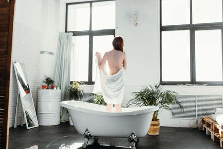 Rear shot of slim young red-haired female model standing in bathtub against big windows, using a towel to hide her nudityの写真素材