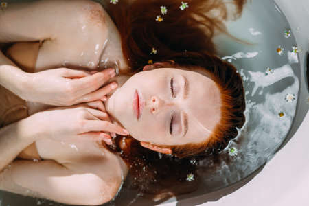 Top view of beautiful sexy red-haired young woman lying in the bathtub with little flowers, eyes closed from pleasure, gently touching her pretty face with healthy skin by hands, close upの写真素材