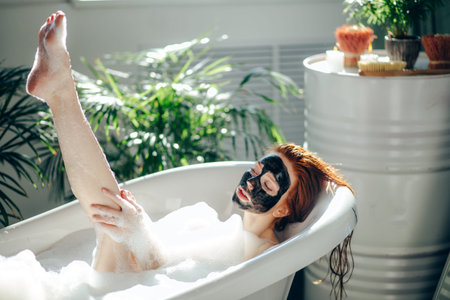 Skin care - Young lady with facial black clay mask applyed on her face relaxing in foam bath,...