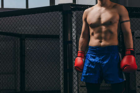 Caucasian young boxer man in red boxing gloves and blue pants stands in dark gym. Concept training boxing.の写真素材