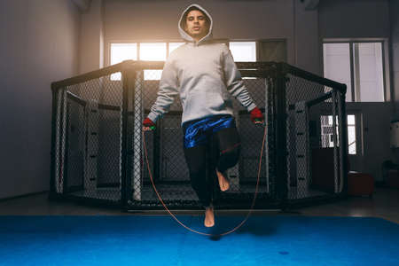 Sportsman in grey hoodie exercises with jump rope near boxing ring, warming up before fightの写真素材