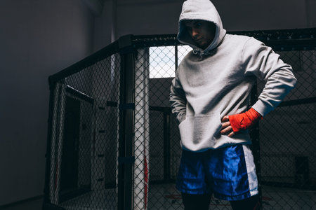 Portrait of male boxer dressed in sports grey hoodie with hood on eyes, blue boxing pants and red bandages on hand posing in fitness studio against chain-link iron mesh ringの写真素材