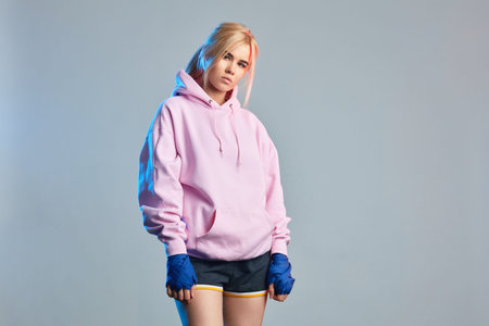 Confident young athlete woman in violet hoodie training in fitness center posing with blue hand...
