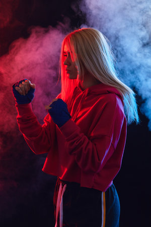 Female blonde boxer in pink hoodie prepares to punch at a boxing studio. Woman boxer in motion over...