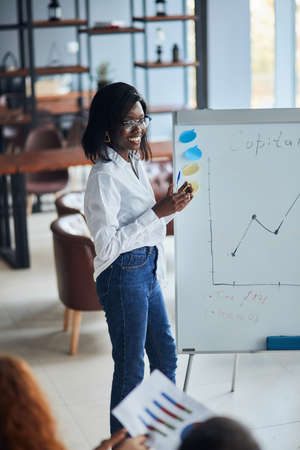 Confident african woman in eyeglasses, wearing white shirt and jeans, using diagrams, motivated african American speaker coach present business plan on whiteboard for workers in modern officeの写真素材