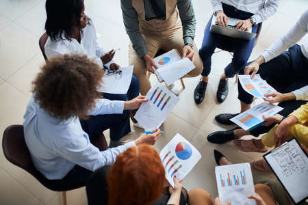 Modern and enthusiastic business people sit in circle discussing business ideas, steps for building business, using diagrams and papers.の写真素材