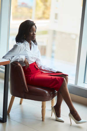 Dark-skinned female business professional sitting at a specious and airy office with large panoramic windows. Corporate employee of a business company dressed in smart casual outfit.の写真素材