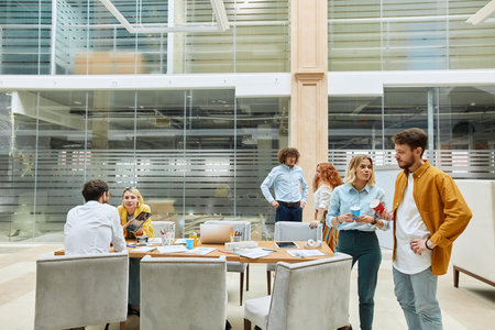 Talented young colleagues stand in pairs chatting with each others during breaktime in roomy,...