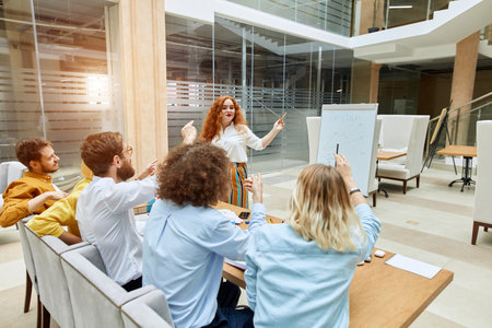 Charming attractive lady stands in front of curious team, points at white paper desk, holds pencil,...