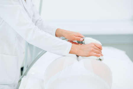 Close up shot of therapist beautician makes LPG massage treatment to young woman in white transparent suit, lying at beauty SPA clinic. Focus on hands holding medical unit.の写真素材