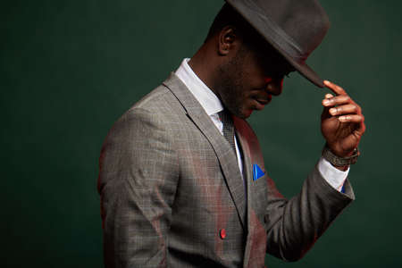 Business and career concept. Portrait of young successful dark-skinned entrepreneur wearing elegant suit and black hat, posing in profile isolated in dark green studioの写真素材