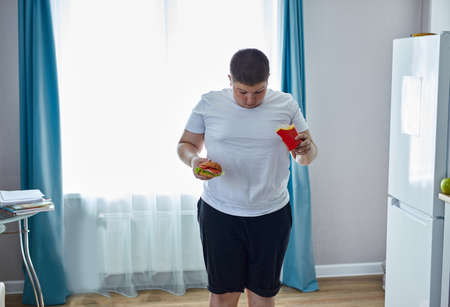 fat teen boy check his weight on scales, want to lose some kilo but at the same time he is eating unhealthy junk food. at homeの写真素材