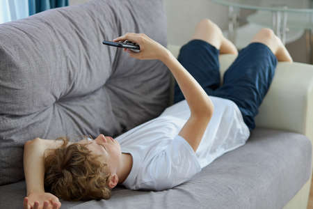 young redhead teen boy watching interesting videos on mobile phone, caucasian curly boy on sofa in living room, have restの写真素材